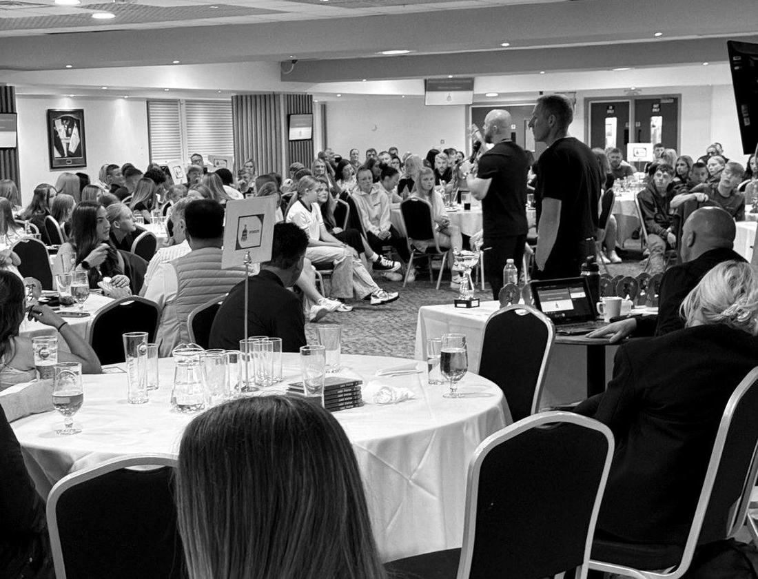 Luke Staton speaking to a full room at a schools football awards evening, copies of Credo stacked on the table.